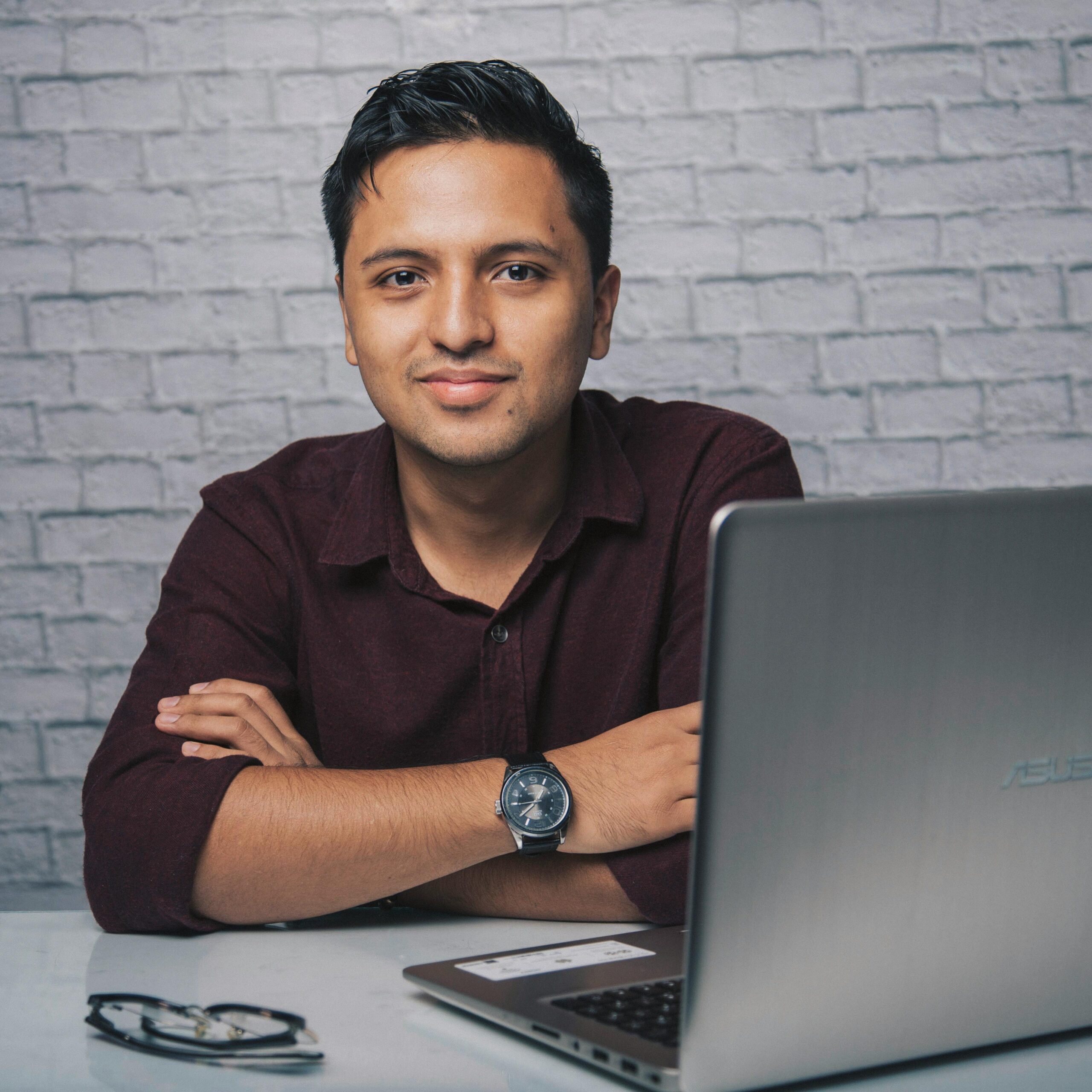 Positive Indian male sitting at table with laptop and eyeglasses with crossed arms while smiling and looking at camera in light office